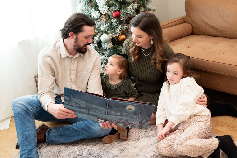 A family shares a joyful moment while reading together on a rug by their Christmas tree.