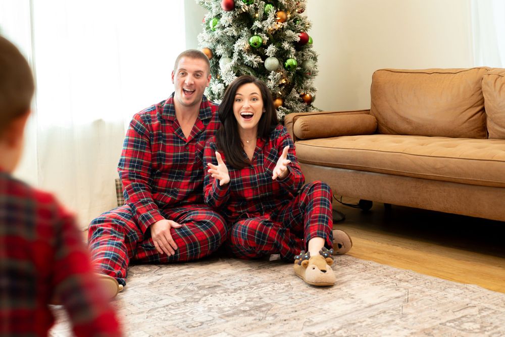 A couple in matching red plaid pajamas laughs together by their decorated Christmas tree.