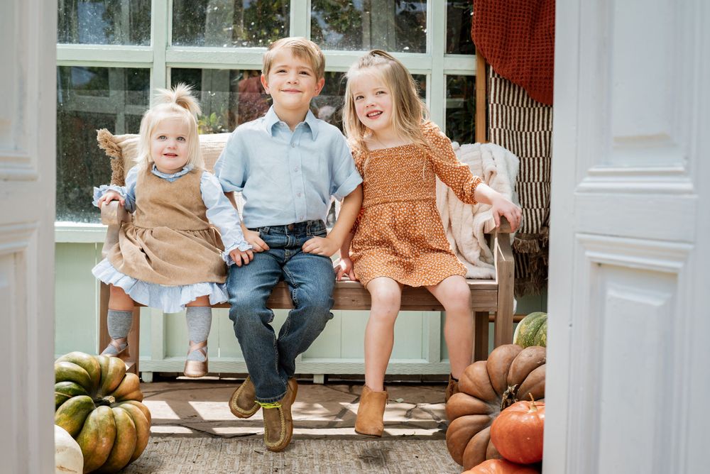 Children sit on a white bench surrounded by autumn pumpkins and gourds.
