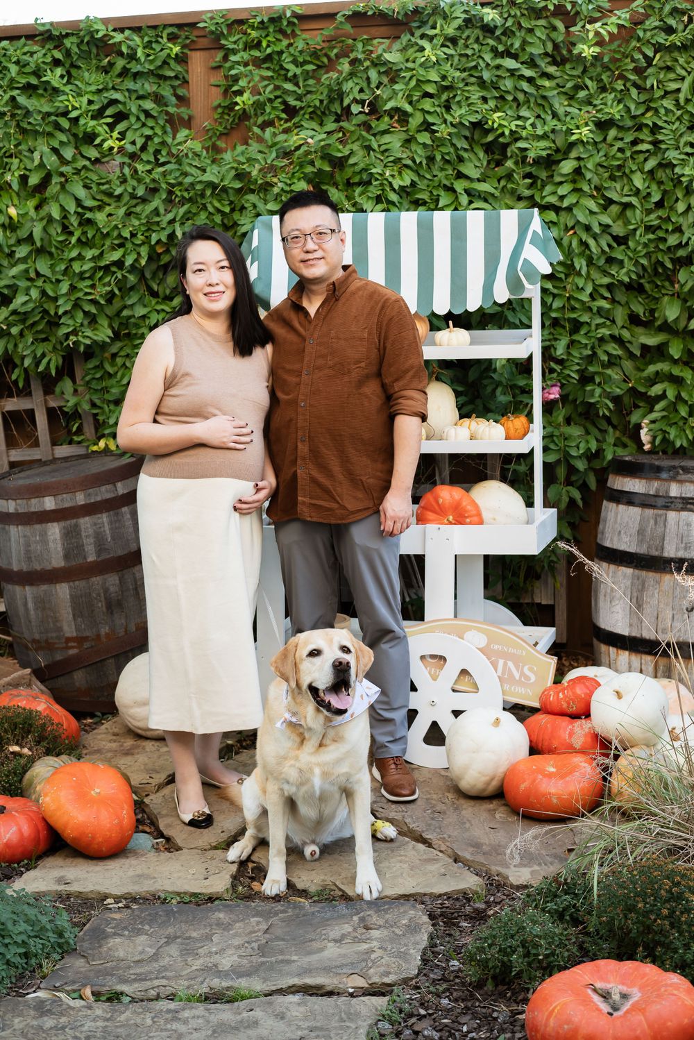 A couple with their golden retriever dog poses among fall pumpkins and wooden barrels in a garden setting.
