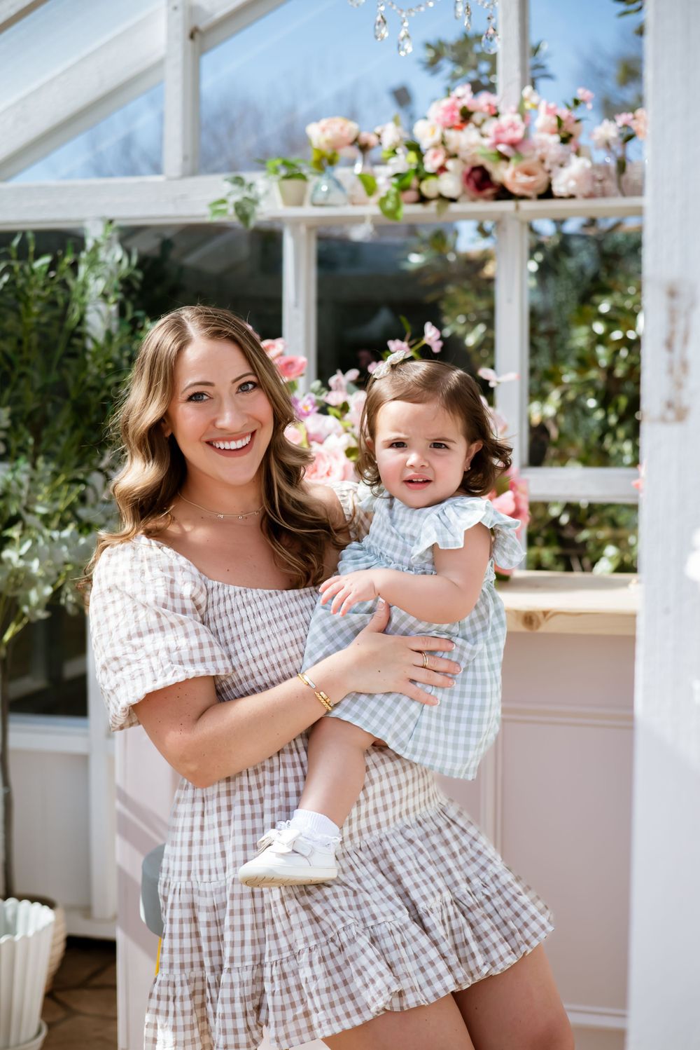 A mother and child pose together in a greenhouse with pink flowers and white decor.