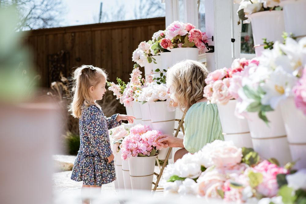 A young girl and woman interact among pink floral arrangements in a bright greenhouse setting.