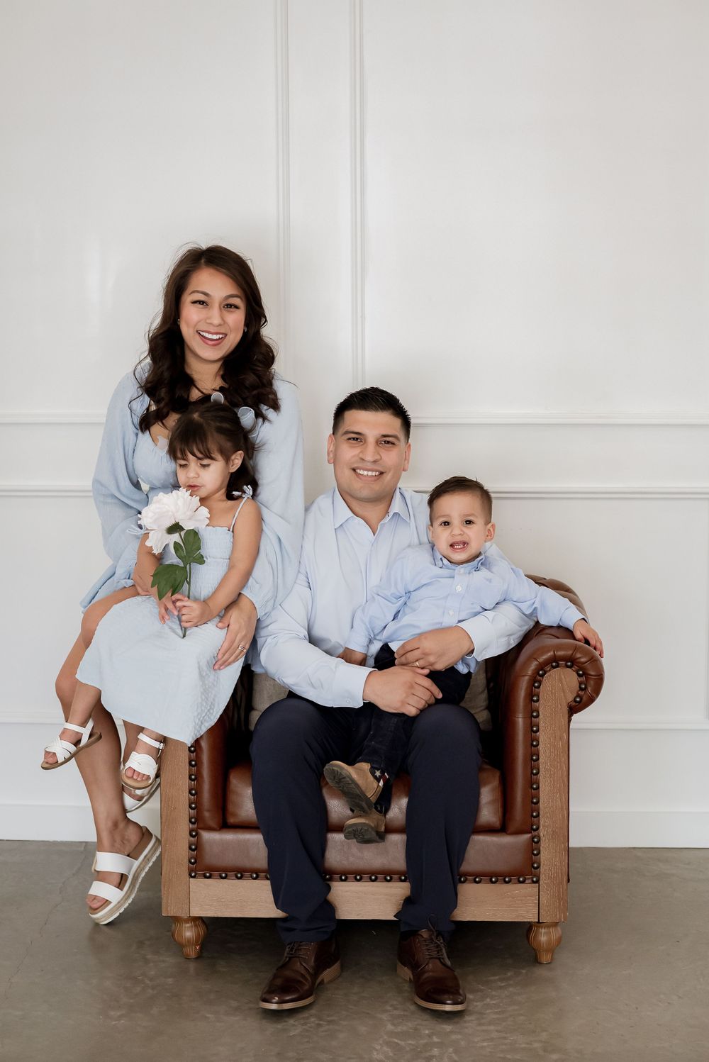 A family of four poses together on a brown leather chair against a white wall in coordinated blue outfits.