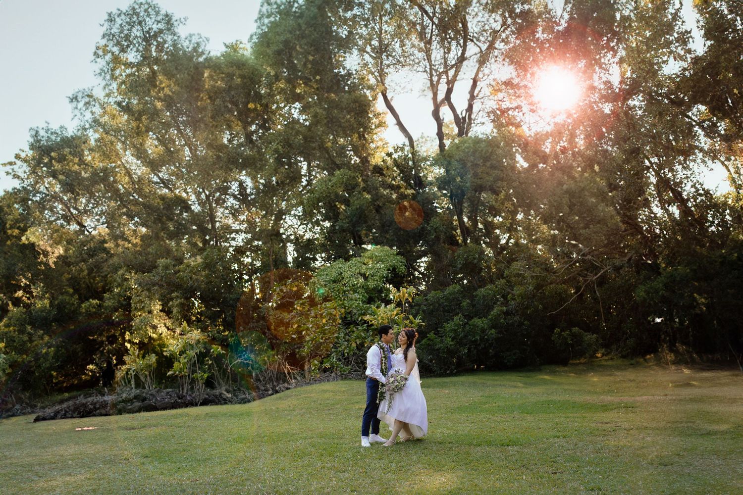 A romantic sunset engagement photo of a couple embracing in a lush garden setting with golden sunlight streaming through trees.