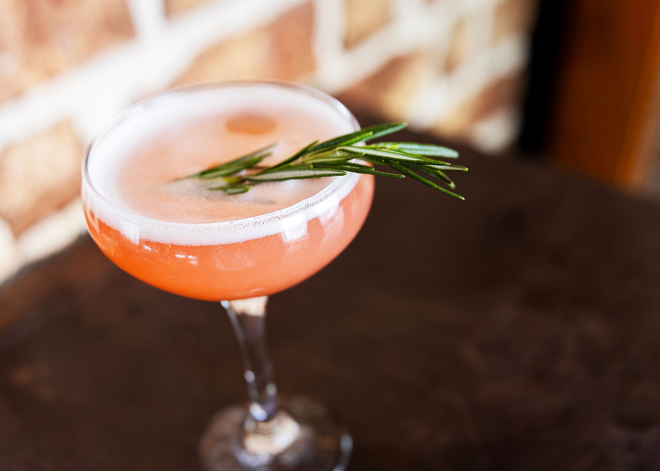 Pink cocktail with rosemary garnish served in a coupe glass on dark wooden surface.