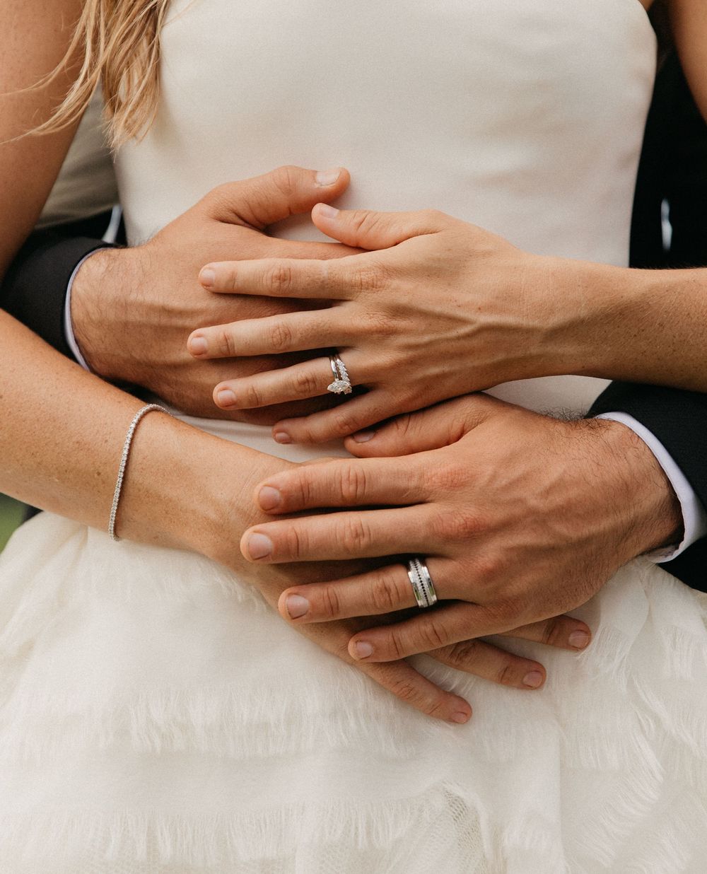 Close-up of bride and groom's hands showing matching wedding bands and diamond engagement ring on white dress.
