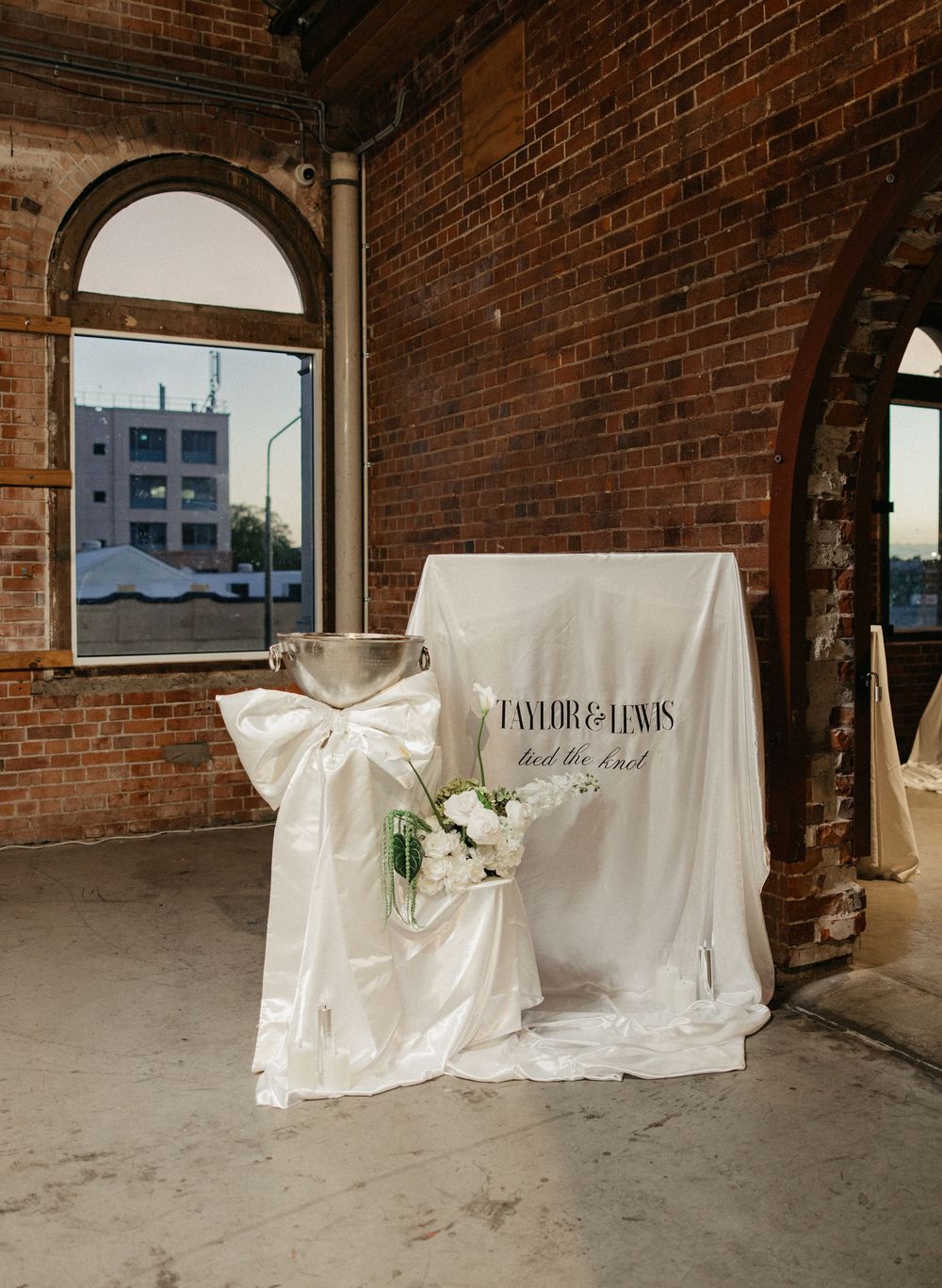Elegant wedding signage display with white satin bow and floral arrangement in a rustic brick venue.