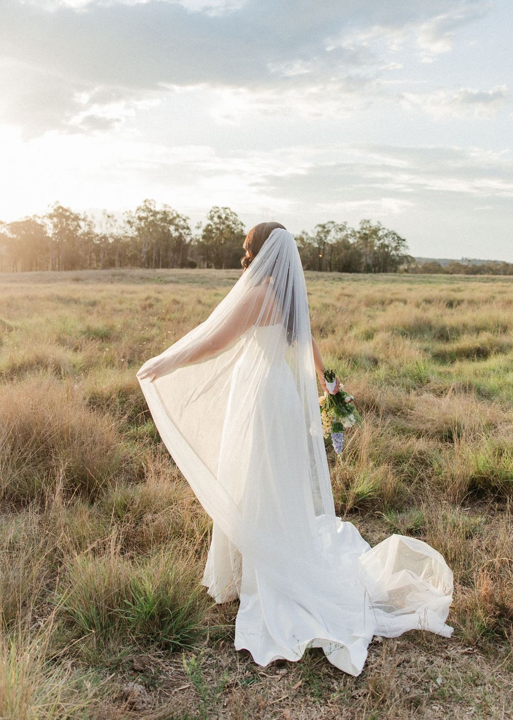 Bride in white gown and flowing veil stands in golden field at sunset holding bouquet.