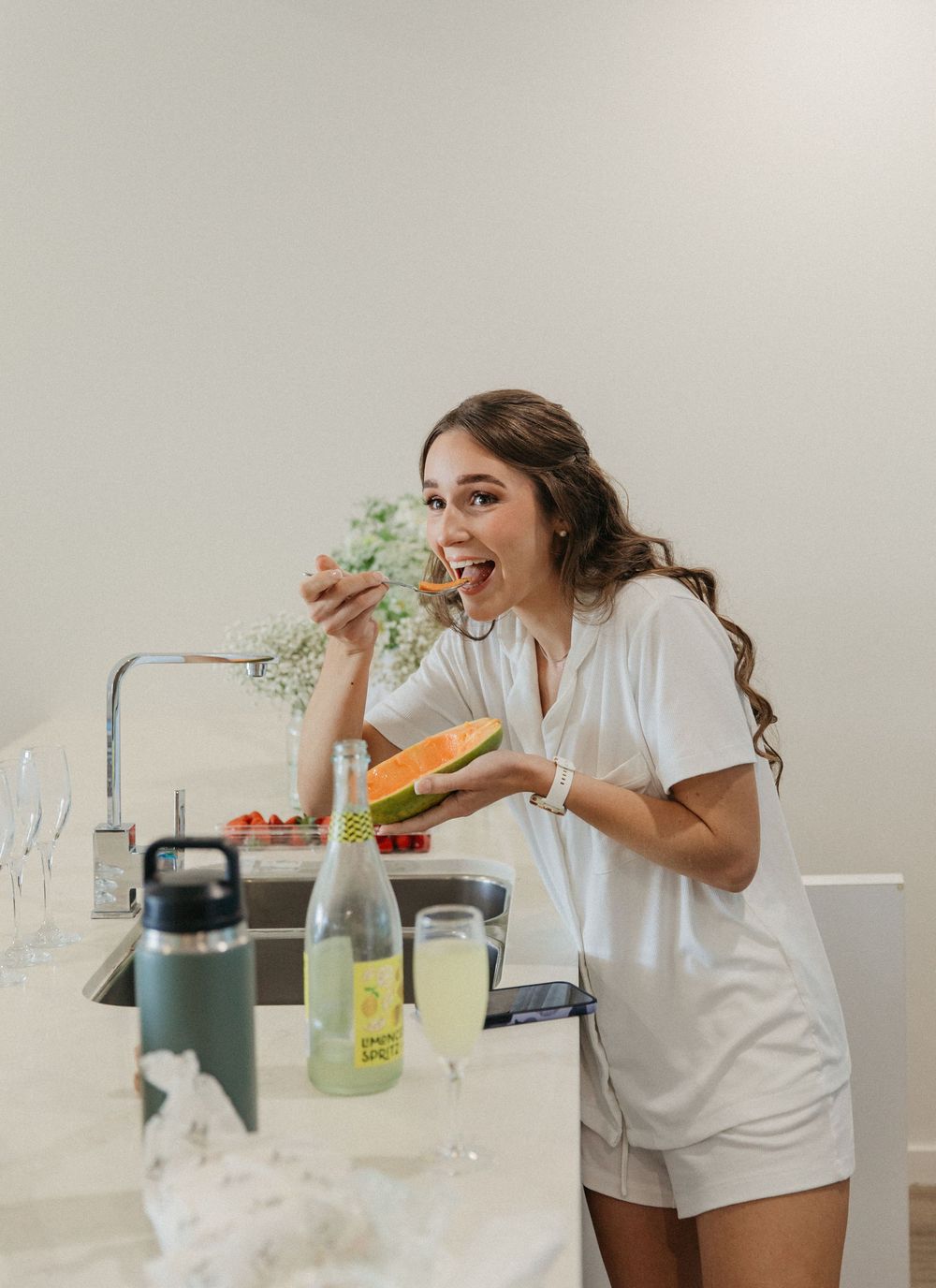 Bride in white romper laughing while eating melon during wedding morning preparations indoors.