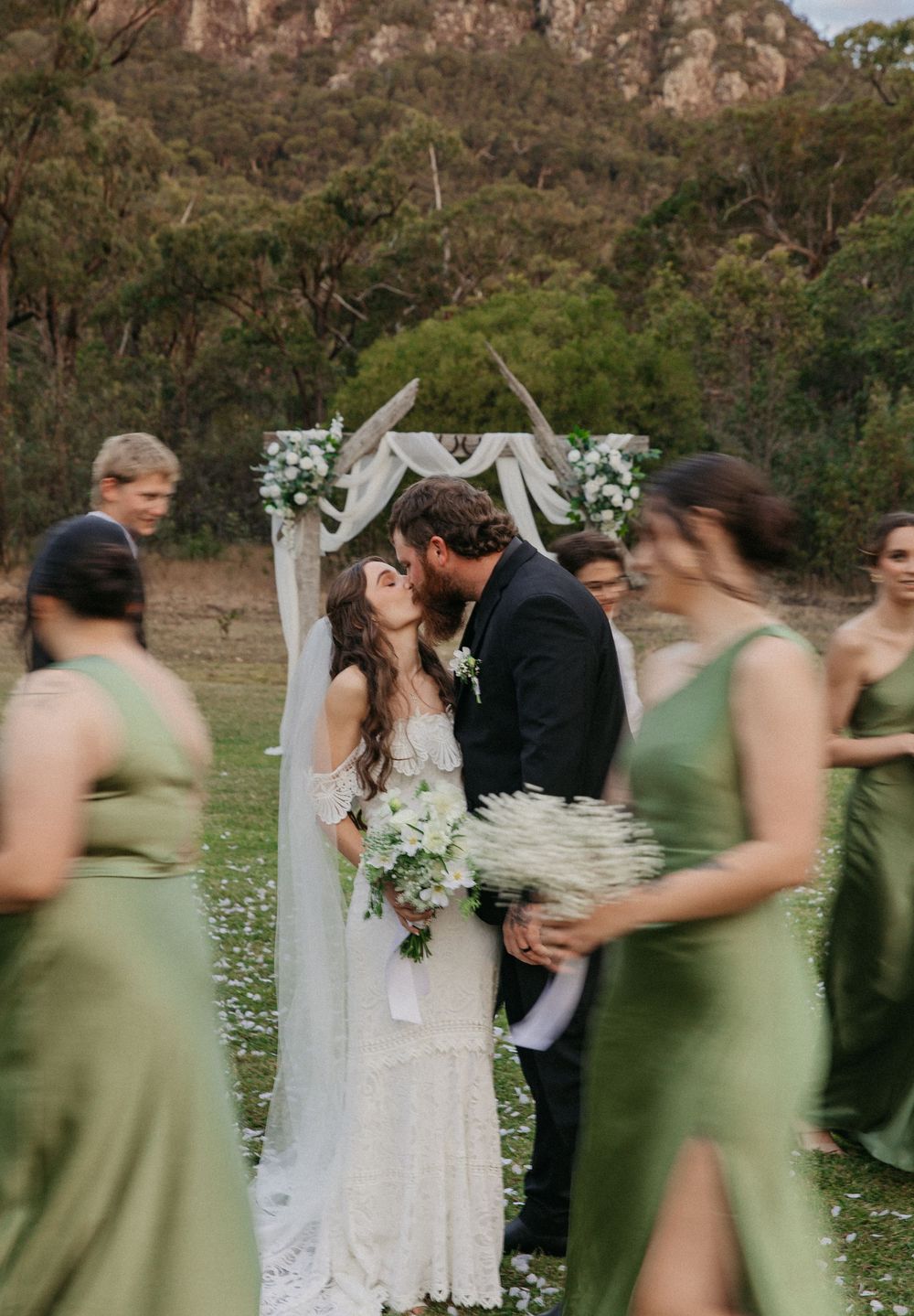 Bride and groom share first kiss under rustic arch with bridesmaids in sage green.