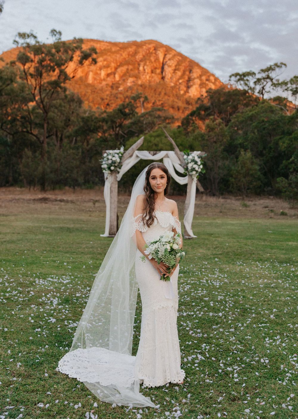 Bride in lace gown and veil stands before rustic arch with mountain at golden hour.