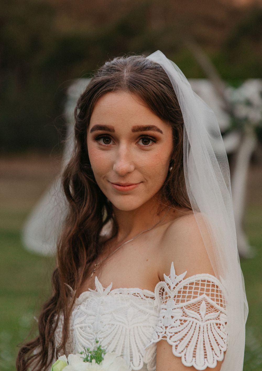 Close-up portrait of brunette bride in off-shoulder lace gown with sheer veil outdoors.