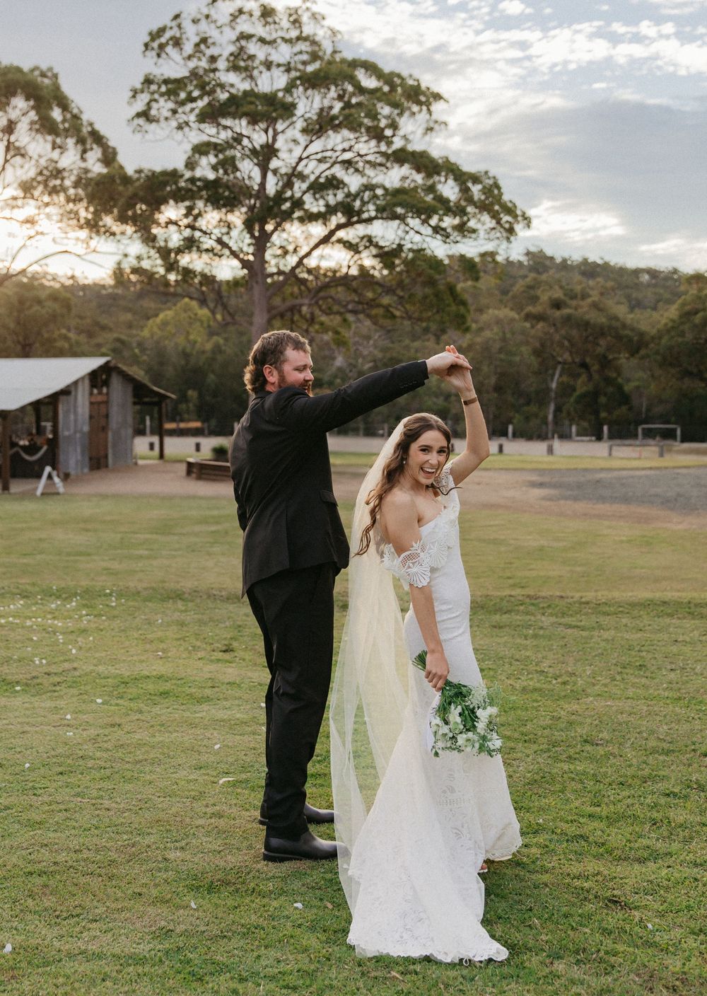 Groom twirling bride in lace wedding dress at golden hour outdoor venue.