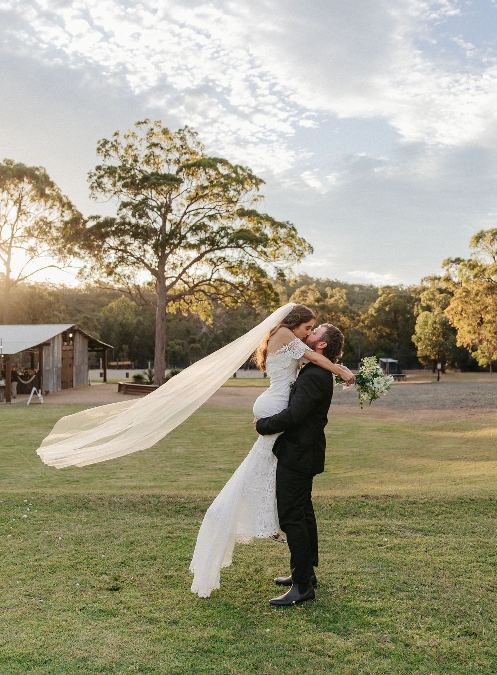 Groom lifts bride for kiss at sunset in scenic outdoor wedding venue.