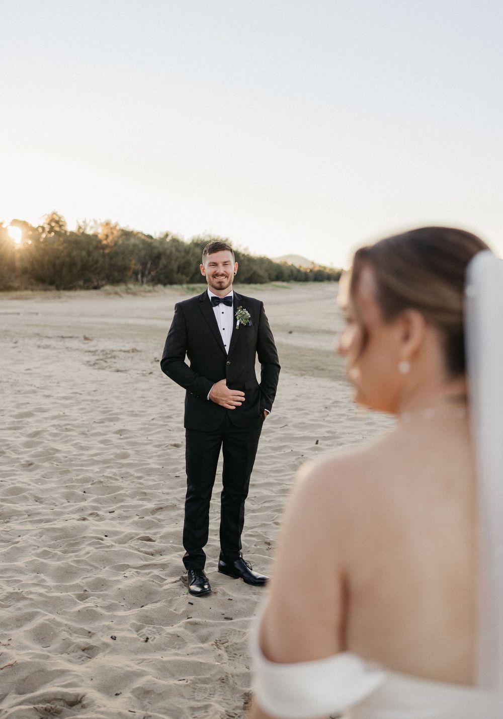 Groom in black tuxedo smiles at bride in white dress during golden hour beach wedding first look.