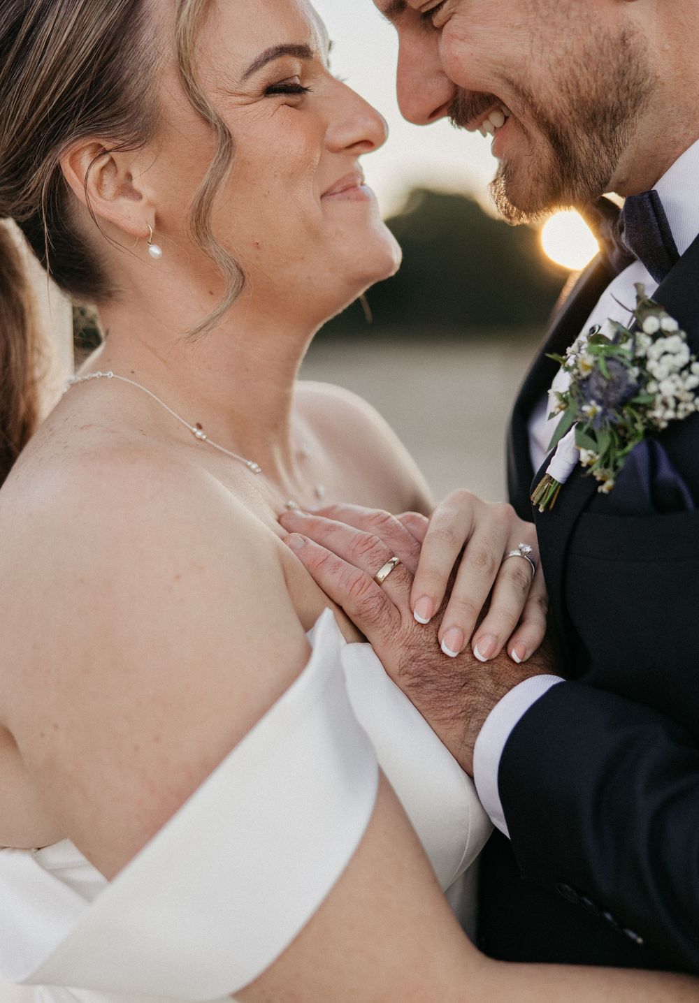 A bride and groom share an intimate moment, holding hands and smiling at each other, wearing wedding rings and formal attire.