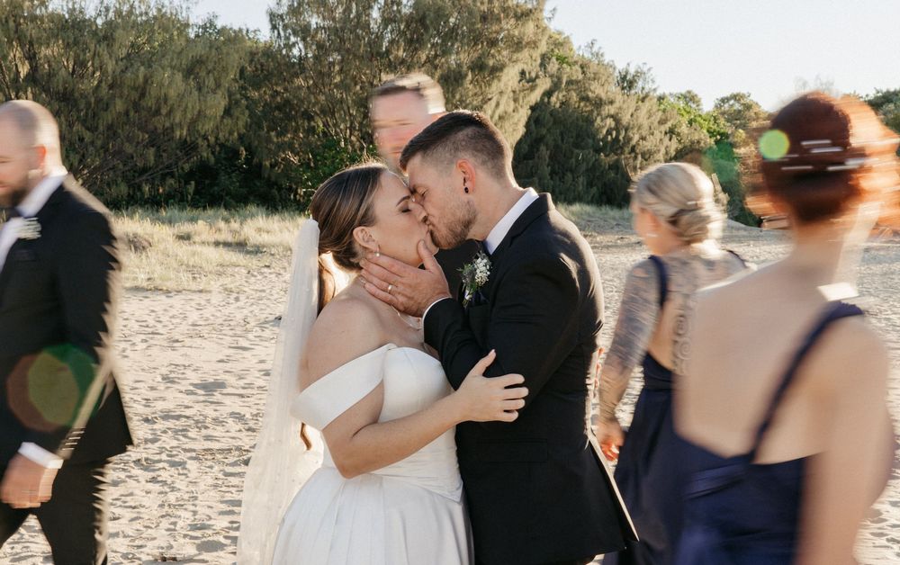 Bride and groom share a romantic kiss on a sandy beach surrounded by wedding guests on their special day.