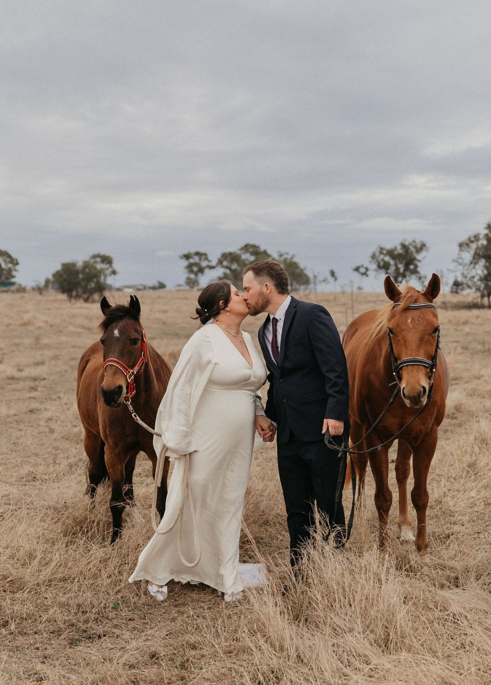 Bride and groom share a kiss between two chestnut horses in a dry grass field.