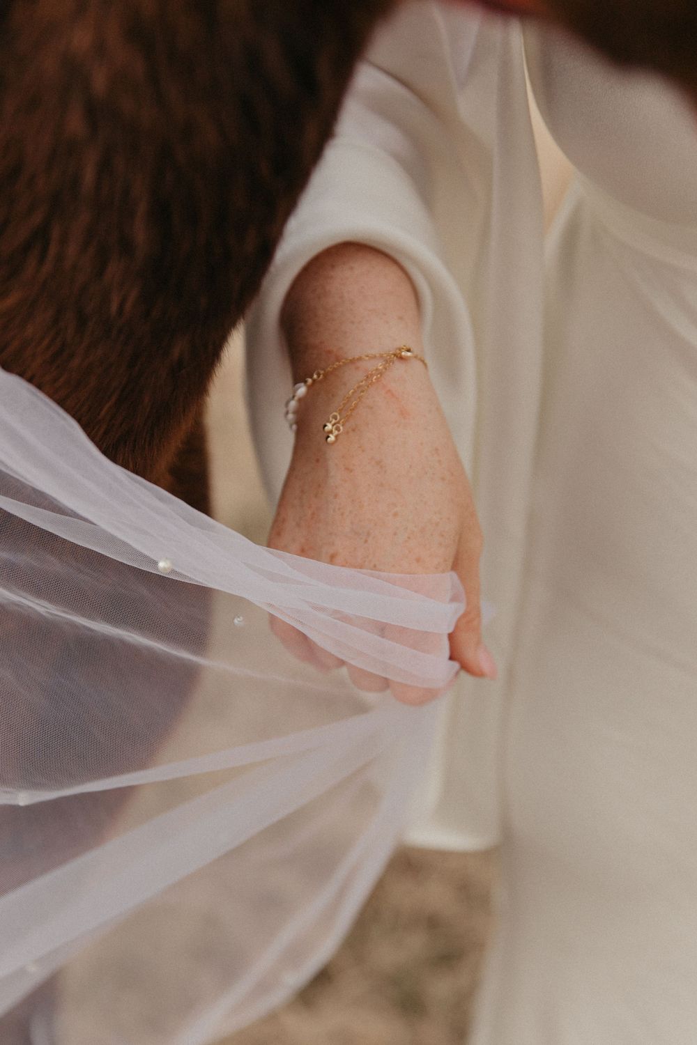Close-up of bride's wrist wearing delicate gold bracelet while holding white veil.