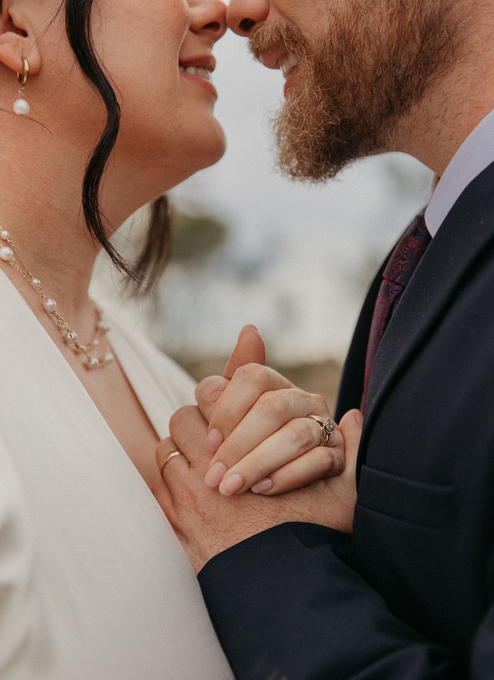 Close-up of bride and groom almost kissing while holding hands showing wedding rings.