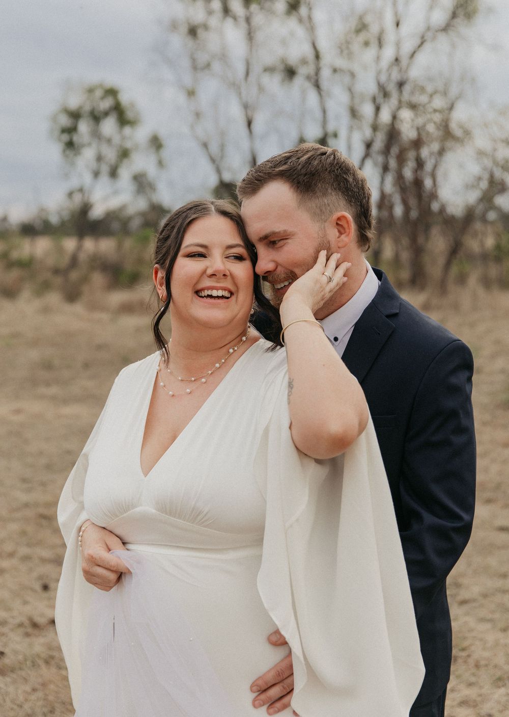 Smiling bride in white cape dress with groom whispering in her ear outdoors.