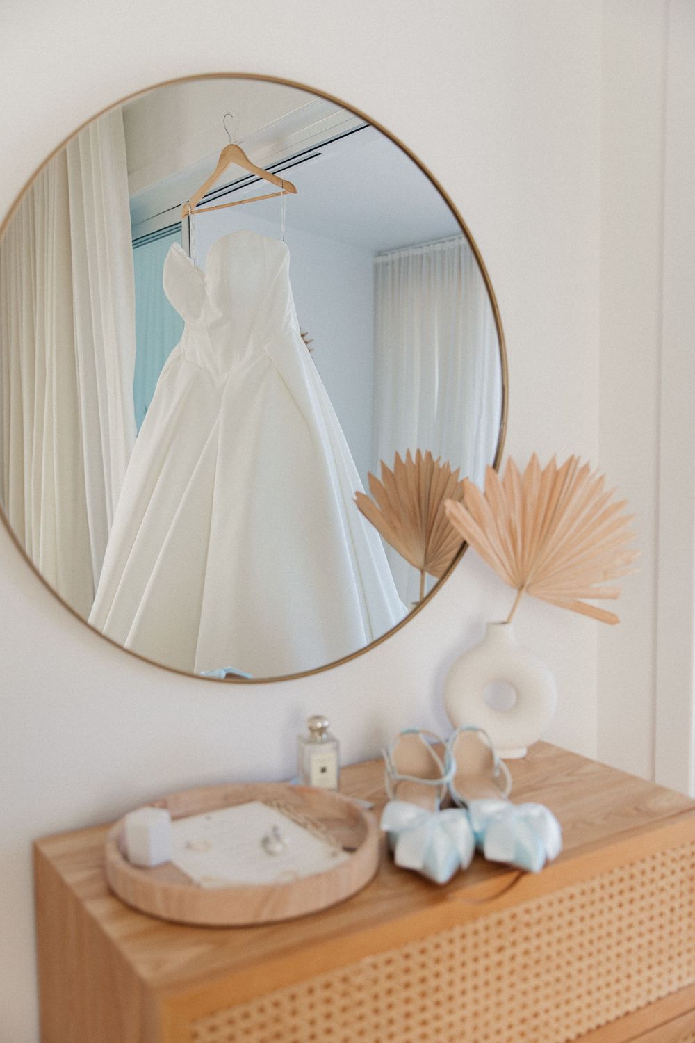 White ballgown wedding dress reflected in round gold mirror above wooden dresser with blue shoes.