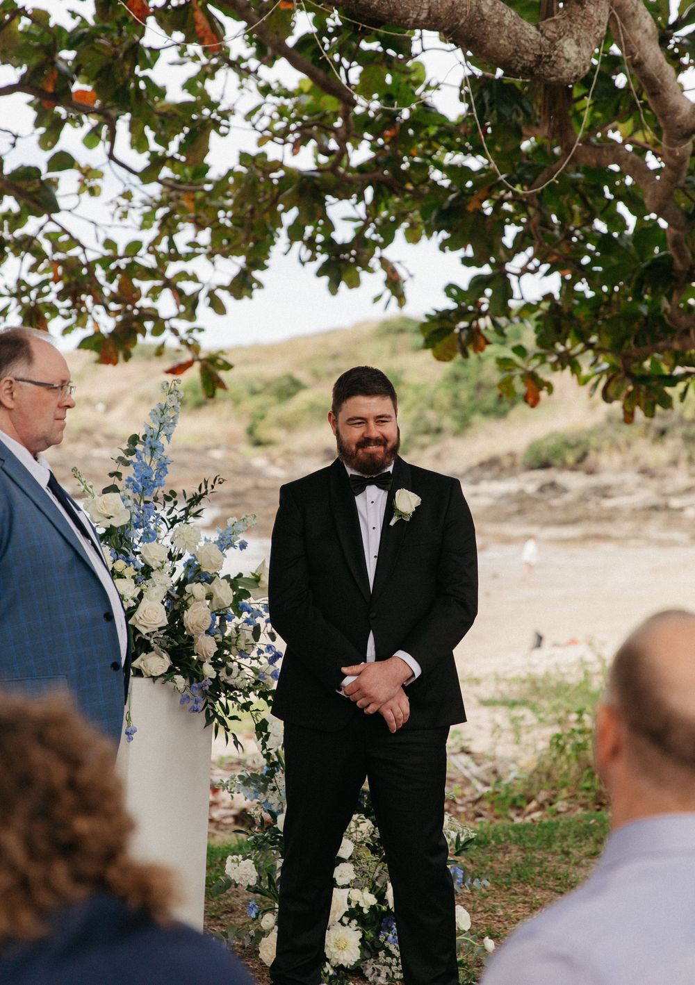 Groom in black tuxedo smiling at altar under tree with white and blue floral arrangements.