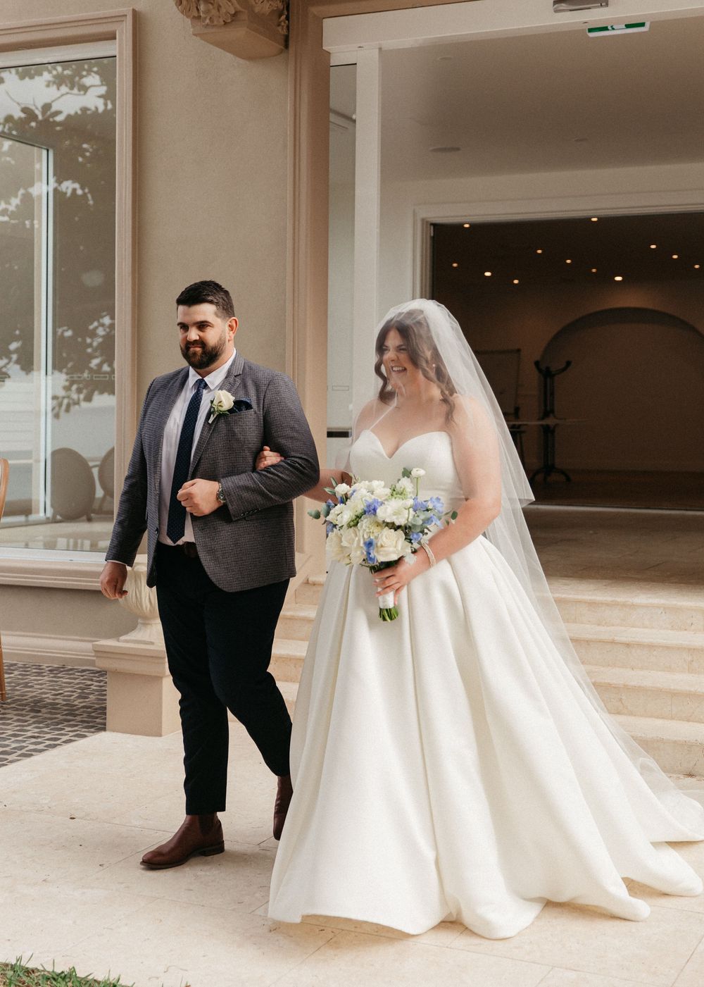 Bride in white ballgown with veil and blue bouquet walking with escort outside wedding venue.