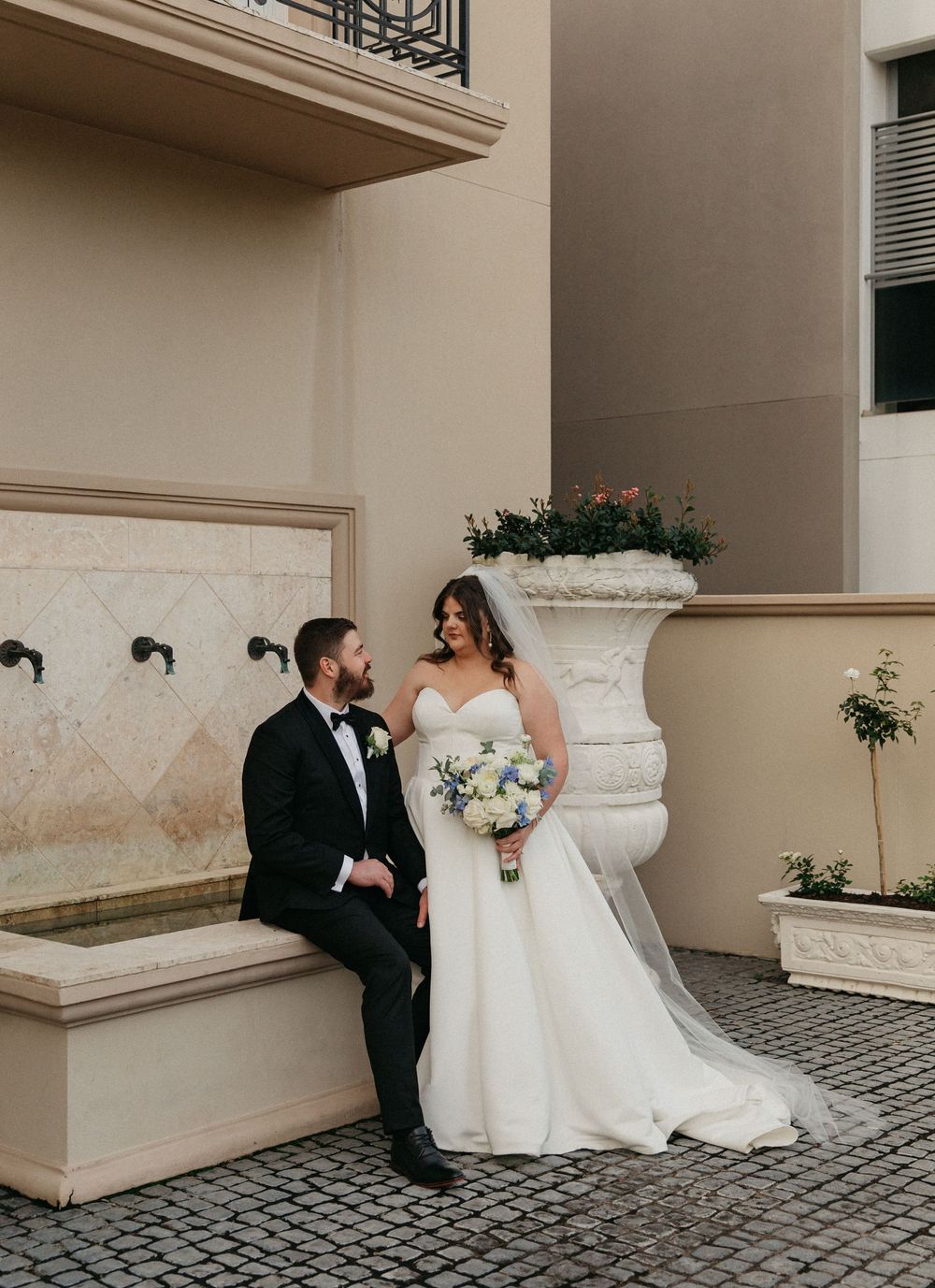 Bride and groom share a romantic moment beside a stone fountain at an elegant venue.
