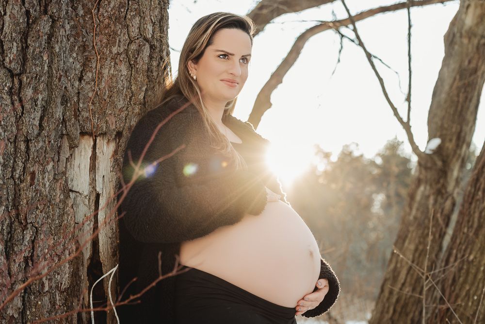 A pregnant woman stands near a tree trunk at sunset with golden sunlight shining through the branches.