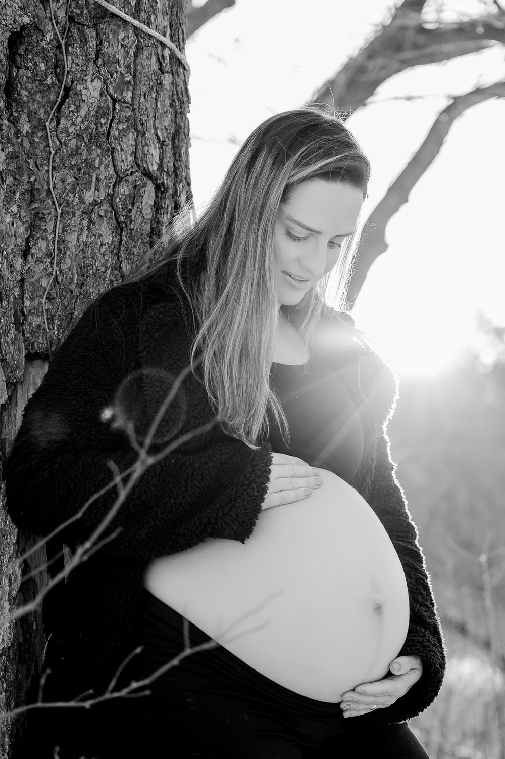 A pregnant woman in a black sweater tenderly cradles her baby bump while posing against a tree in black and white.