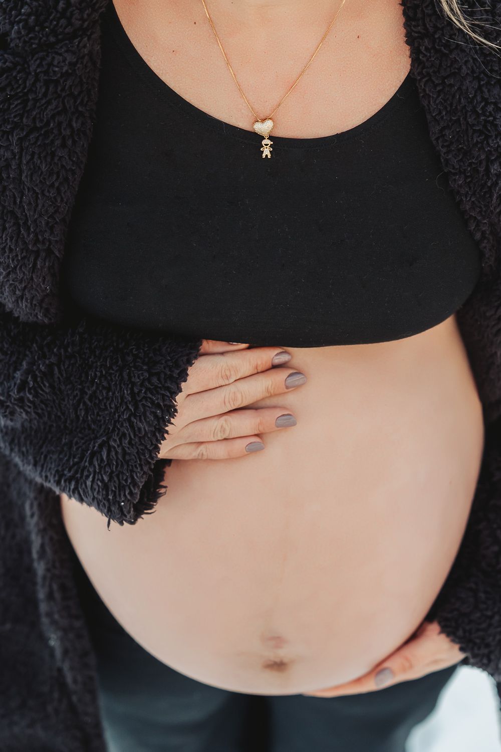 Close-up of pregnant belly with hands resting on bare stomach, wearing black sweater and delicate necklace.