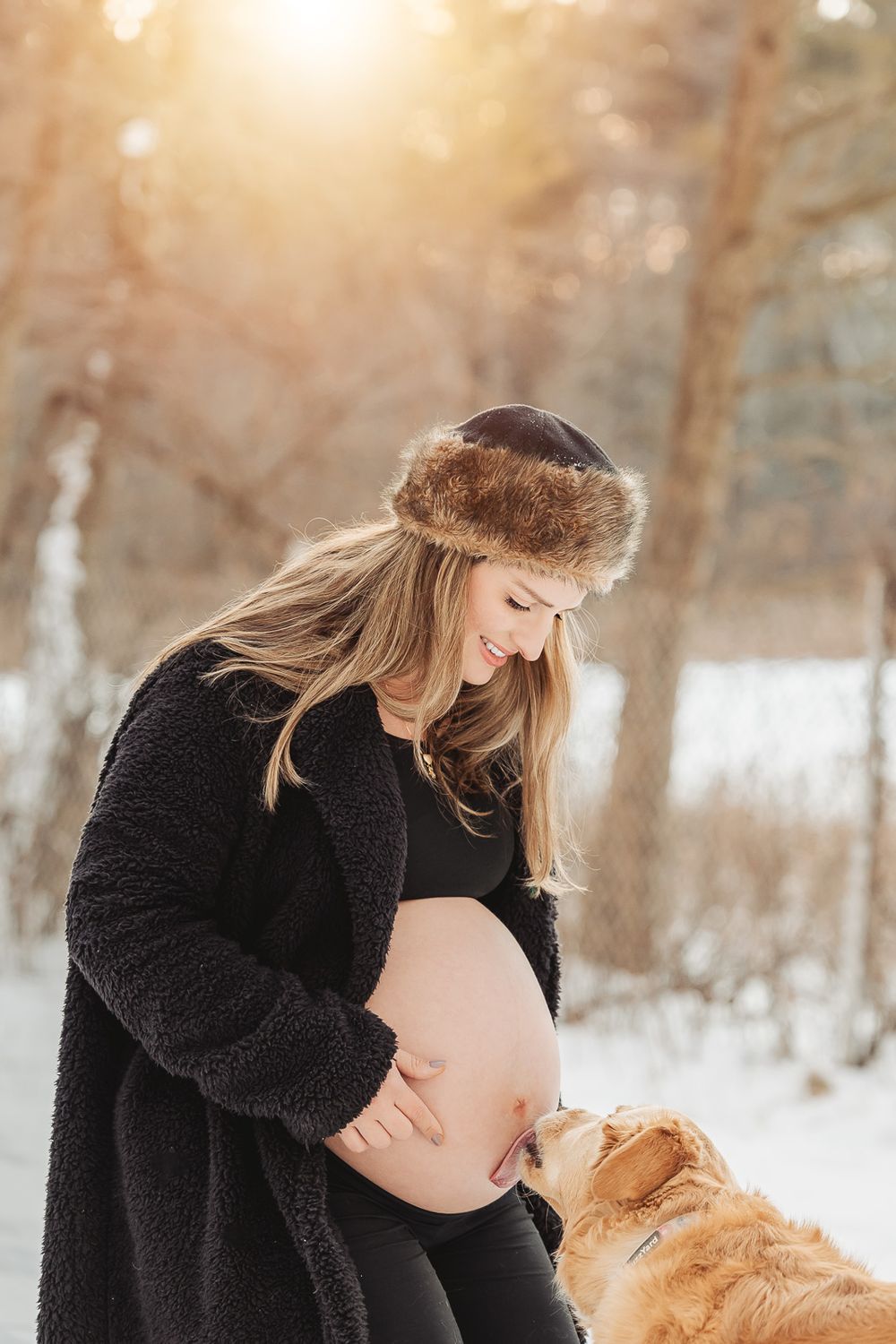 A pregnant woman in winter attire and fur hat shares a tender moment with her golden dog in a snowy forest setting.