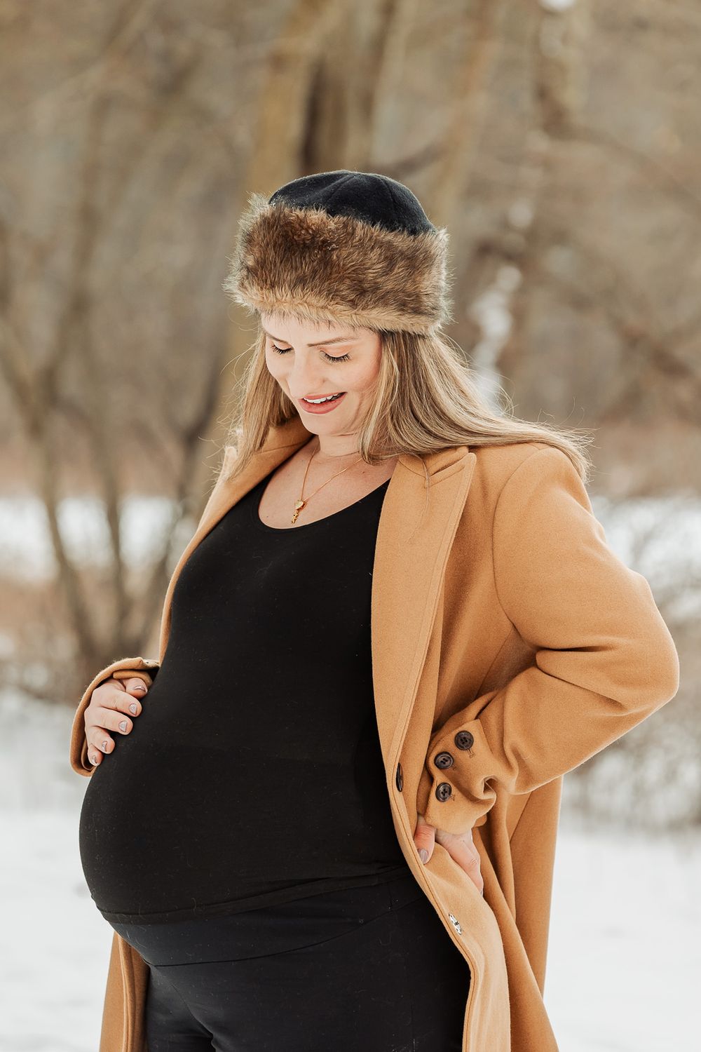 Winter maternity photo with a camel coat and fur-trimmed hat against snowy woods.