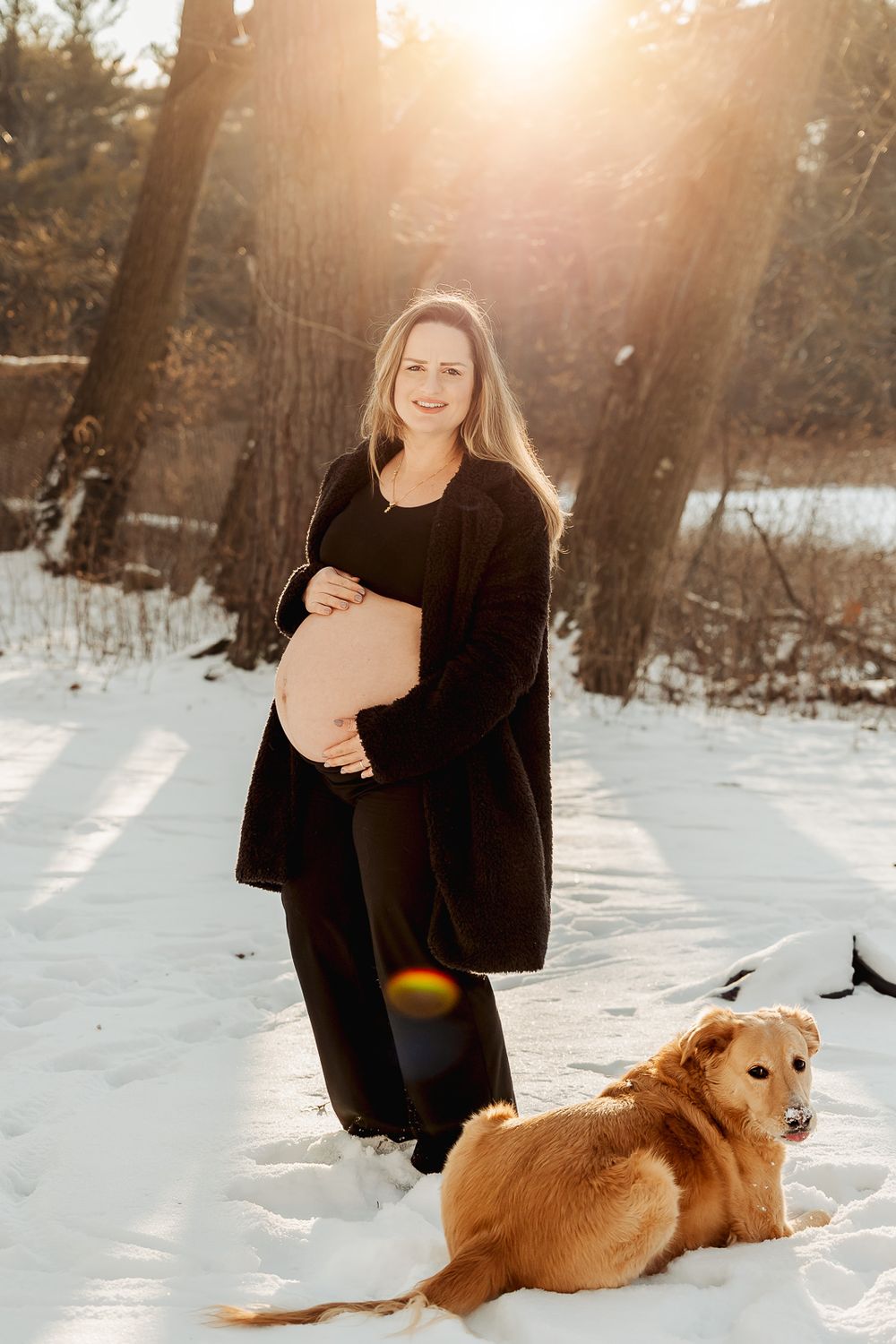 A winter maternity session in a snowy forest with a golden retriever dog lying in the snow at sunset.