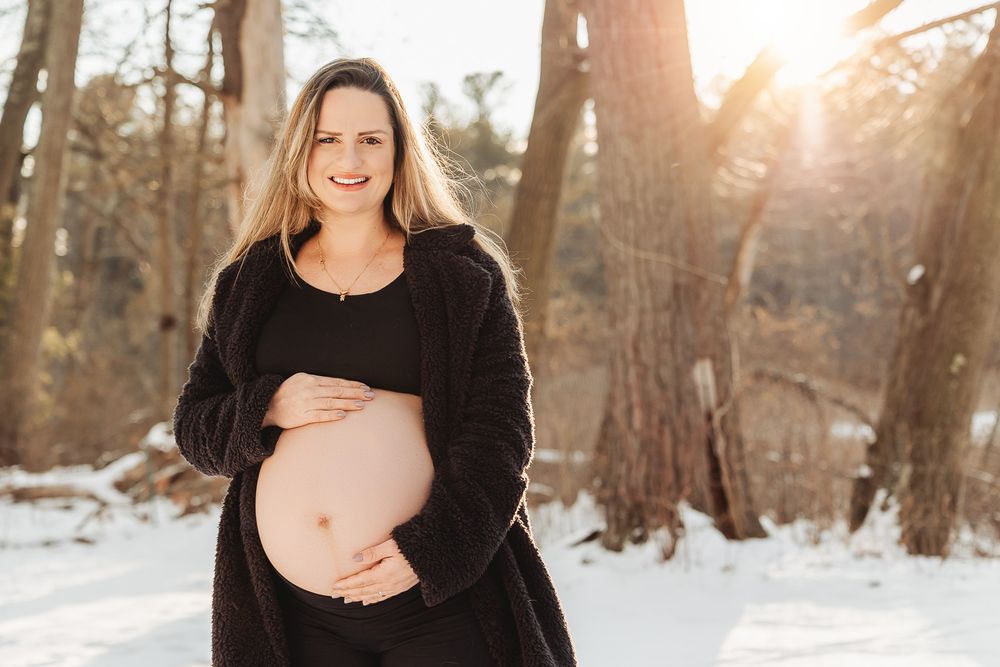 An expectant mother in black clothing stands in a snow-covered forest during golden hour sunset.