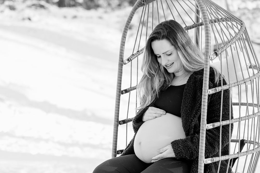 A maternity portrait featuring an expectant mother sitting in a hanging egg chair against a snowy background.