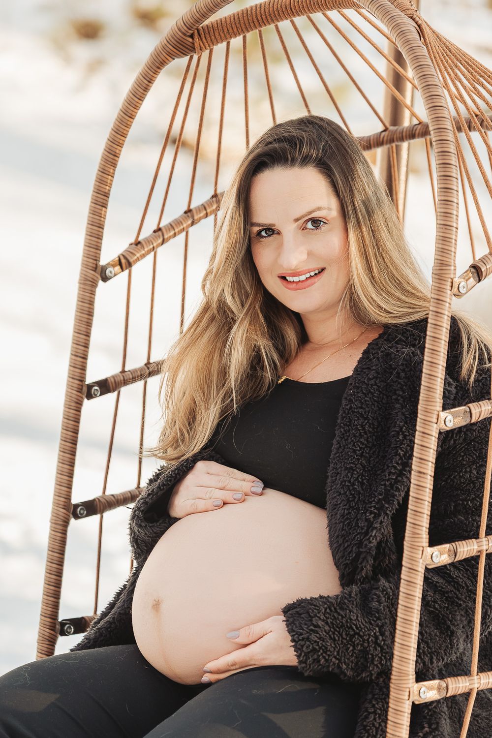 Expectant mother sitting in rattan hanging chair wearing black outfit and cozy cardigan during maternity photoshoot outdoors.