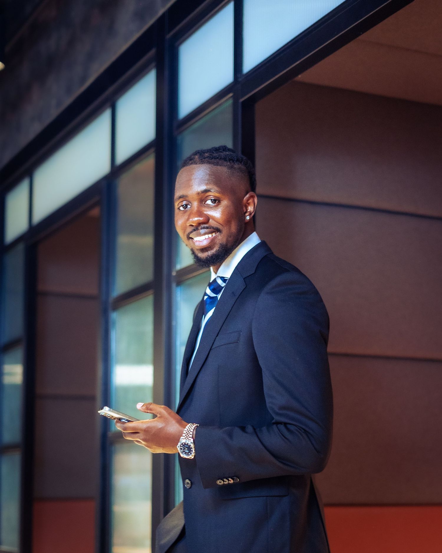 Smiling businessman in a navy suit holding a phone while standing outside a modern building.