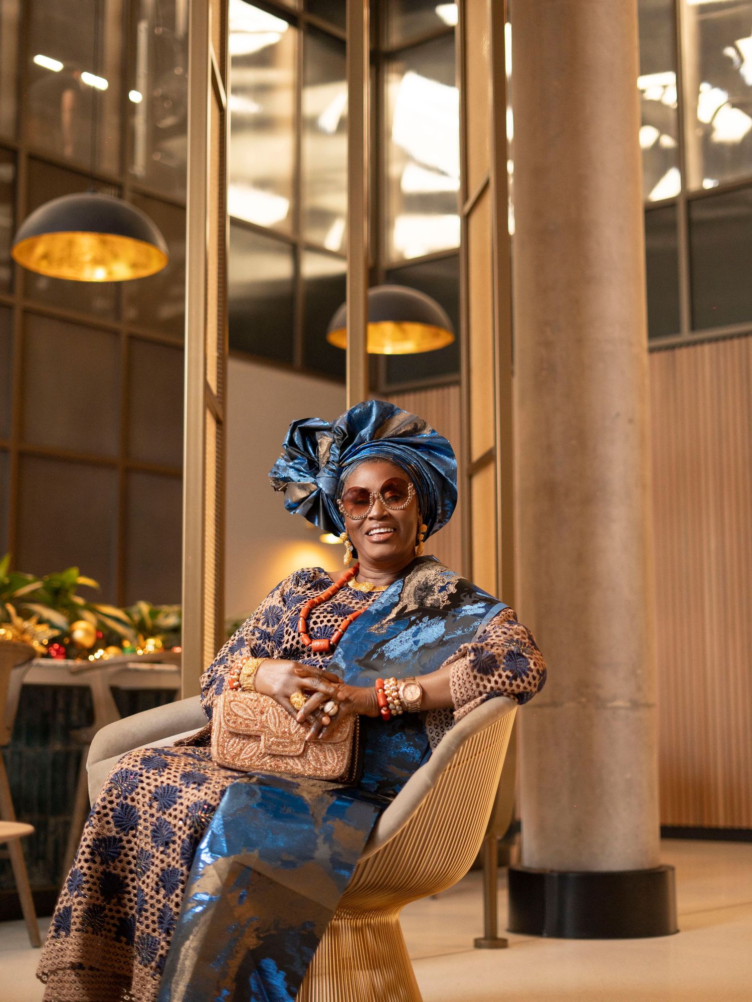 Elegant woman in traditional Nigerian blue ankara outfit and gele headwrap seated in a modern lobby.