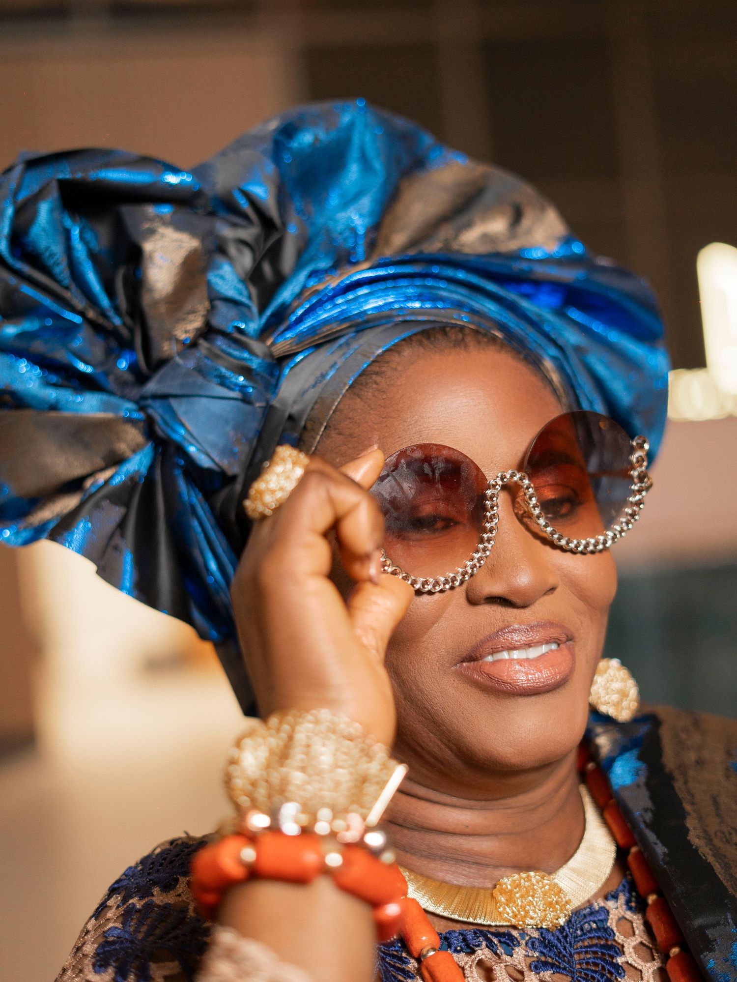 Close-up of a woman in a blue gele headwrap wearing stylish oversized sunglasses and coral beaded jewelry.