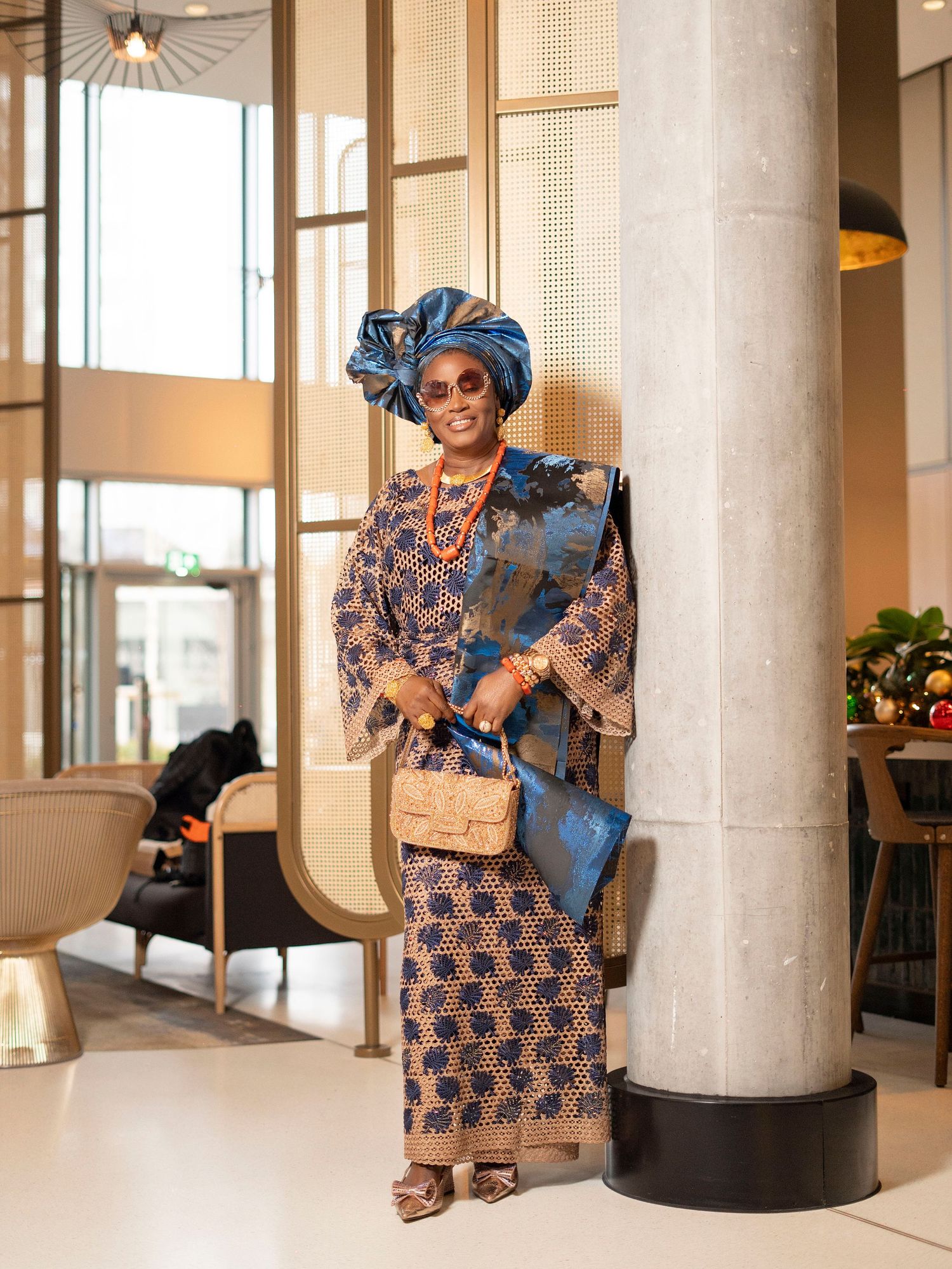 Woman in full traditional Nigerian blue ankara dress and gele standing beside a column in a modern building.