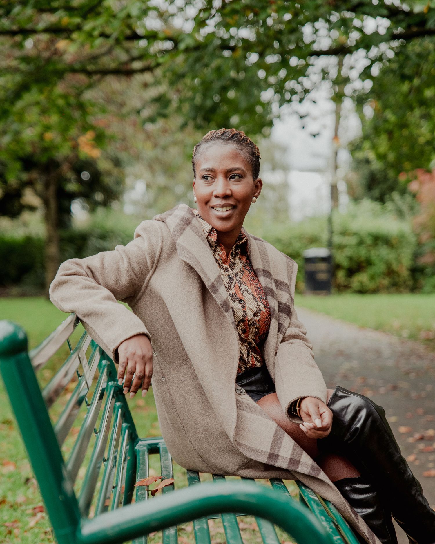 A tall Black woman in a beige coat sits on a green park bench surrounded by autumn foliage.