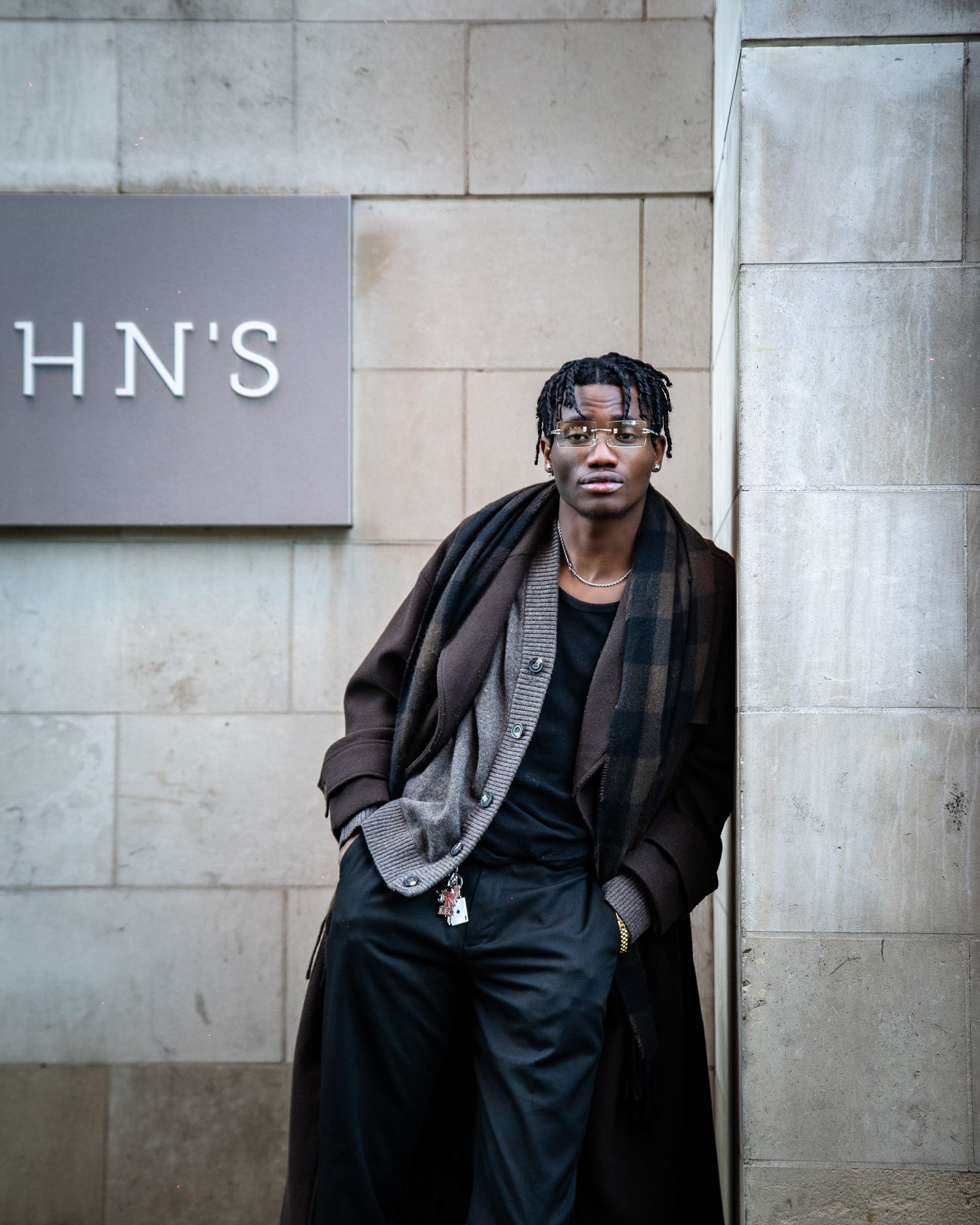Young man in a long plaid coat and black outfit leaning against a stone wall near a sign.
