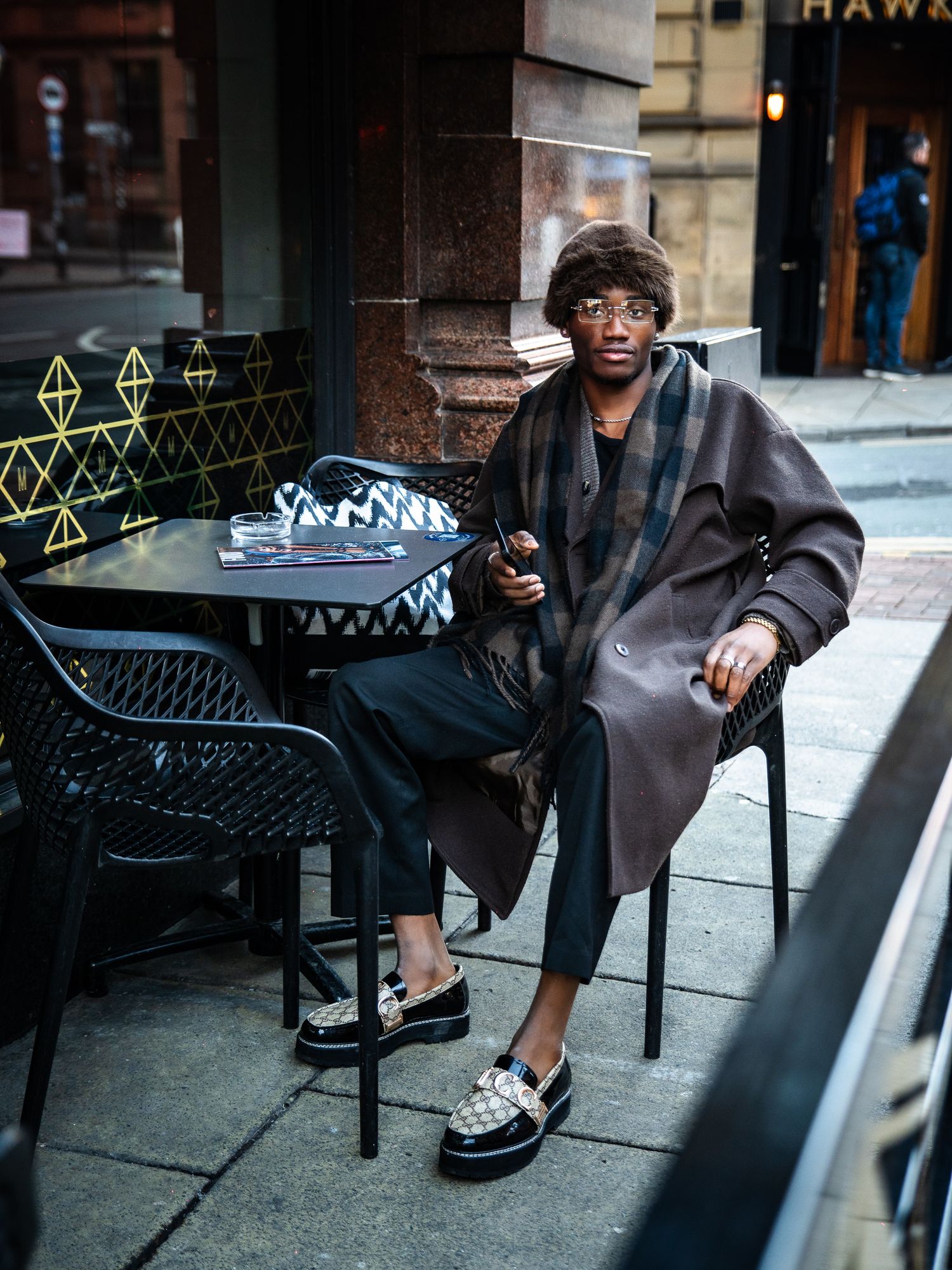 Stylish young man in a plaid coat and loafers sitting at an outdoor cafe table holding a phone.