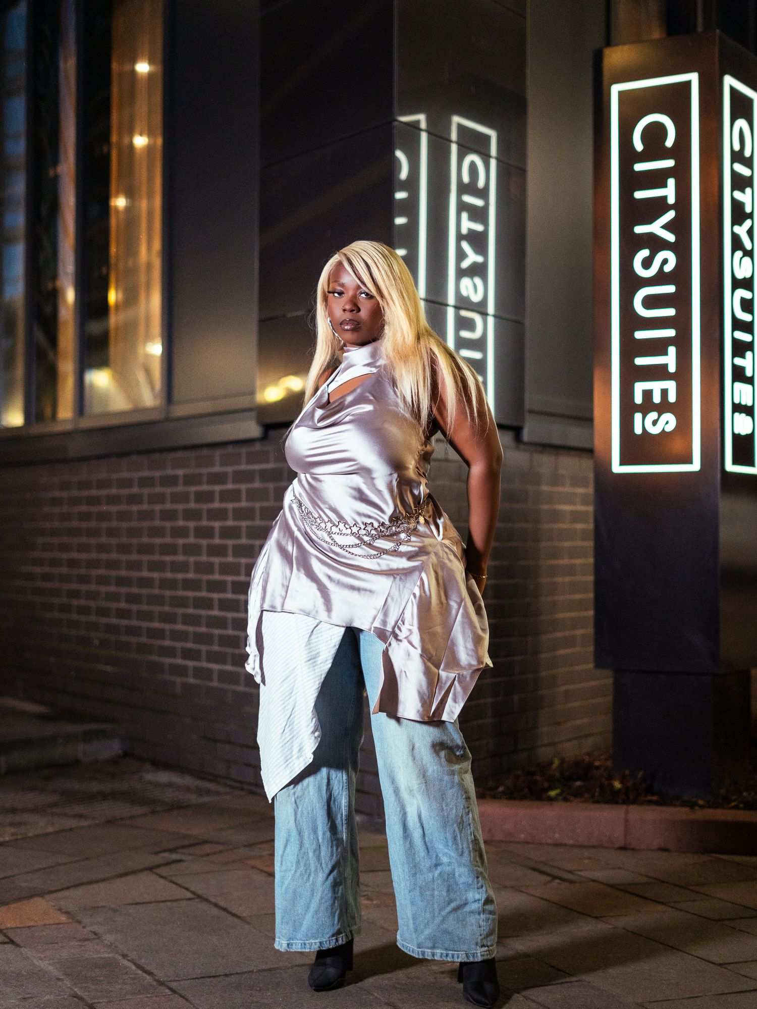 Woman in silver satin top and wide-leg jeans posing at night outside City Suites building.