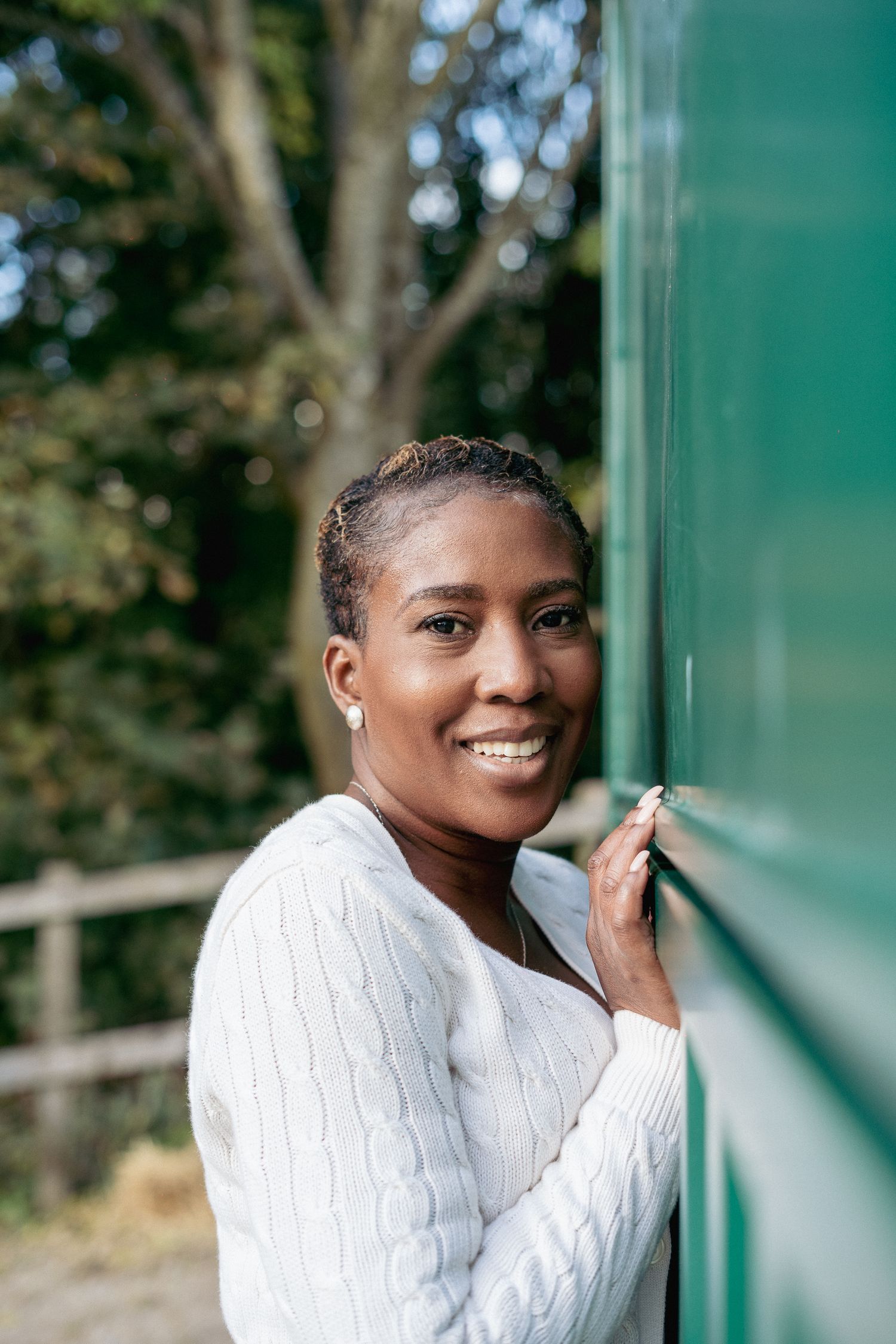 A smiling Black woman with short hair in a white cable-knit sweater leans against a green wall outdoors.