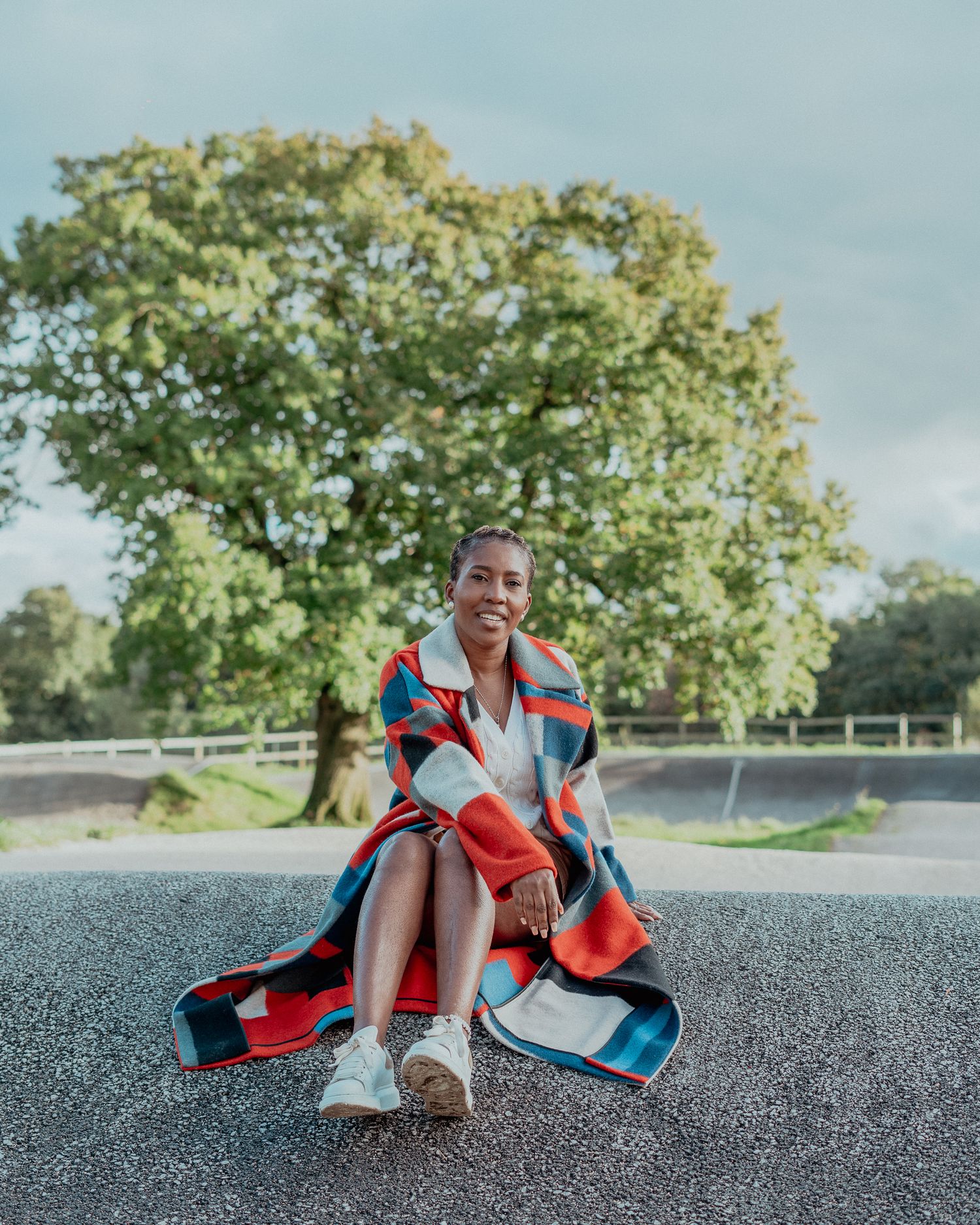 A Black woman in a colorful oversized coat sits on a road with a large green tree in the background.