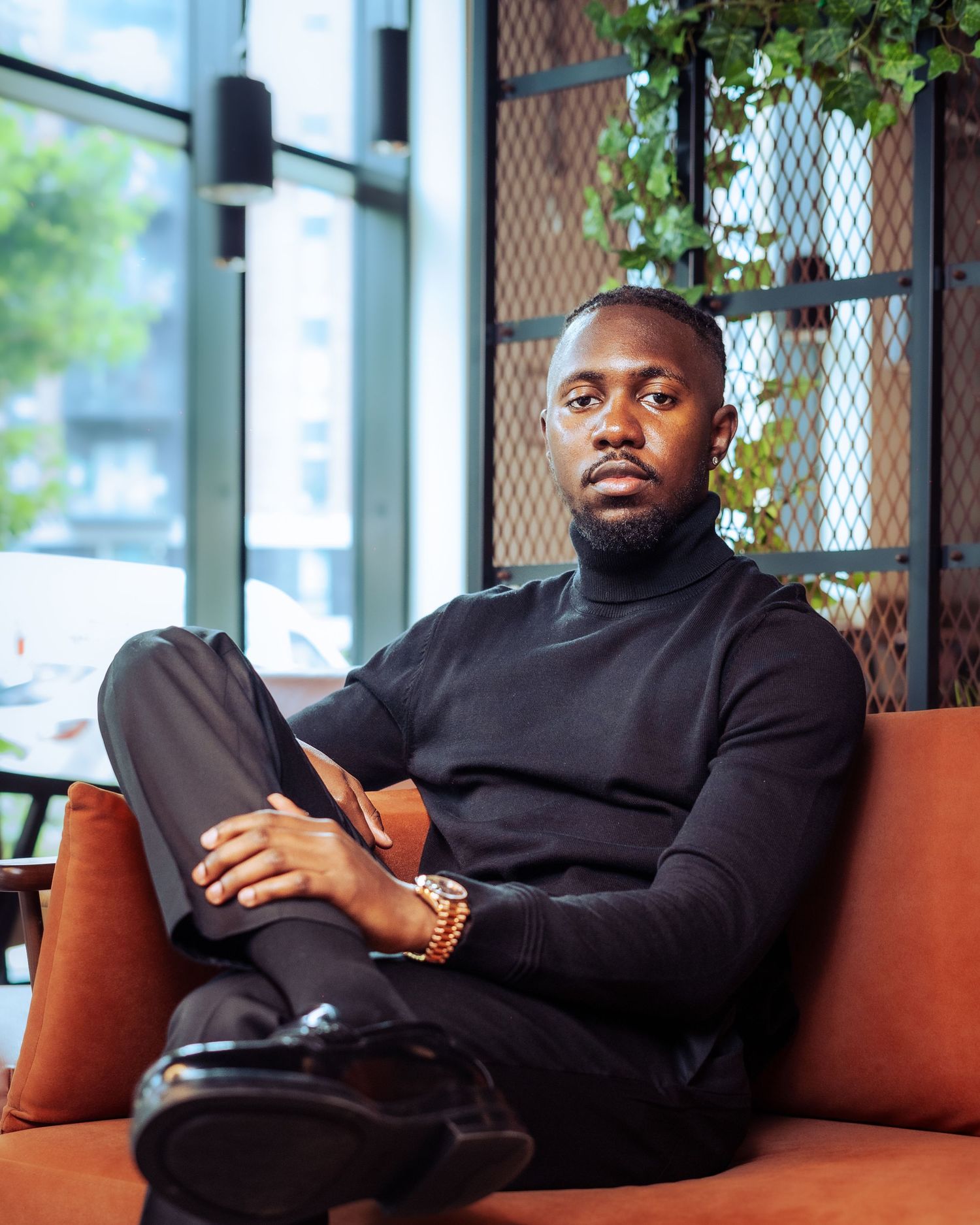 A stylish Black man in a black turtleneck sits relaxed on an orange sofa in a modern cafe setting.