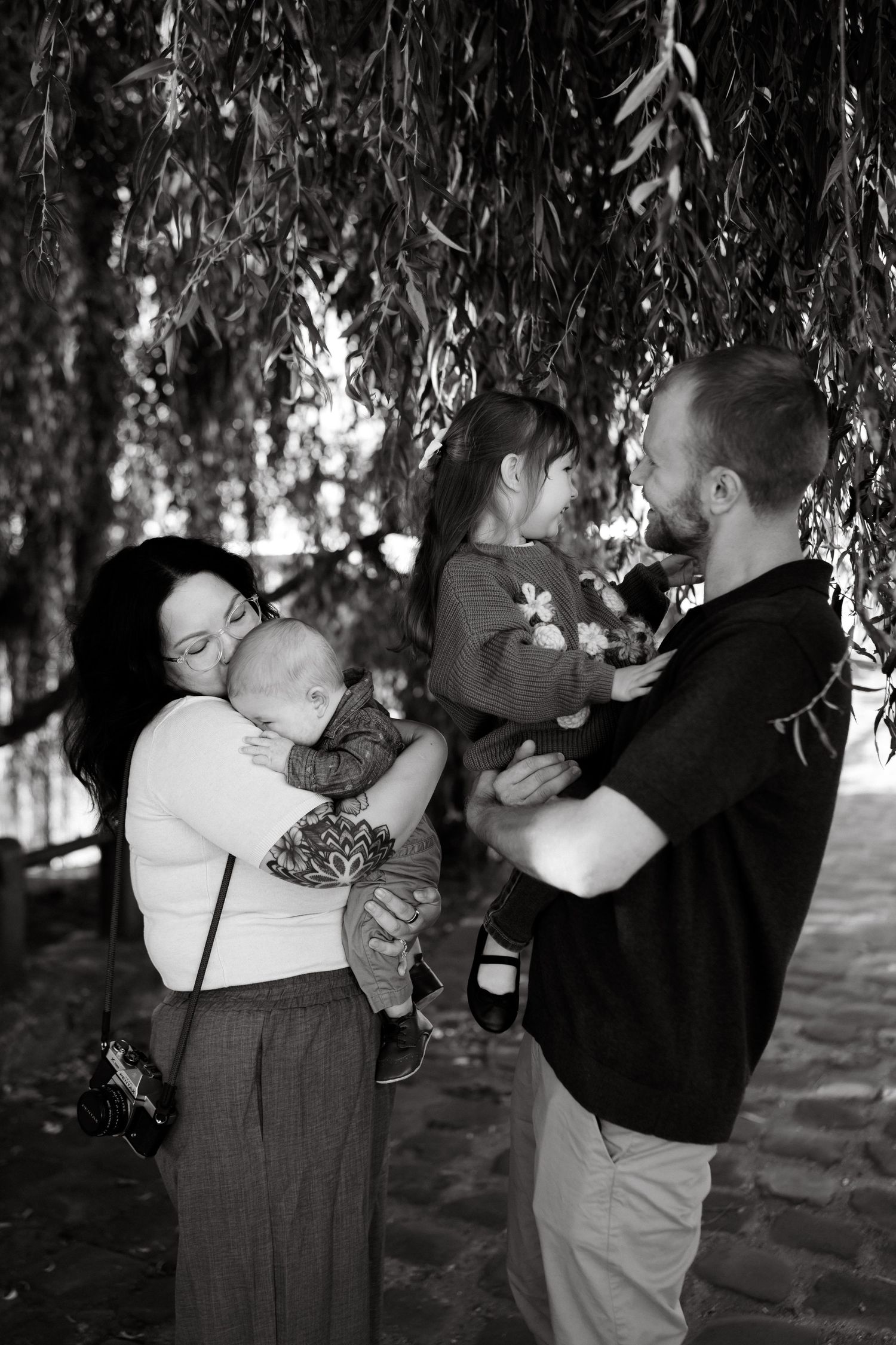 Black and white image of family in Paris France under a willow tree on the Seine River. Mom has a film camera.